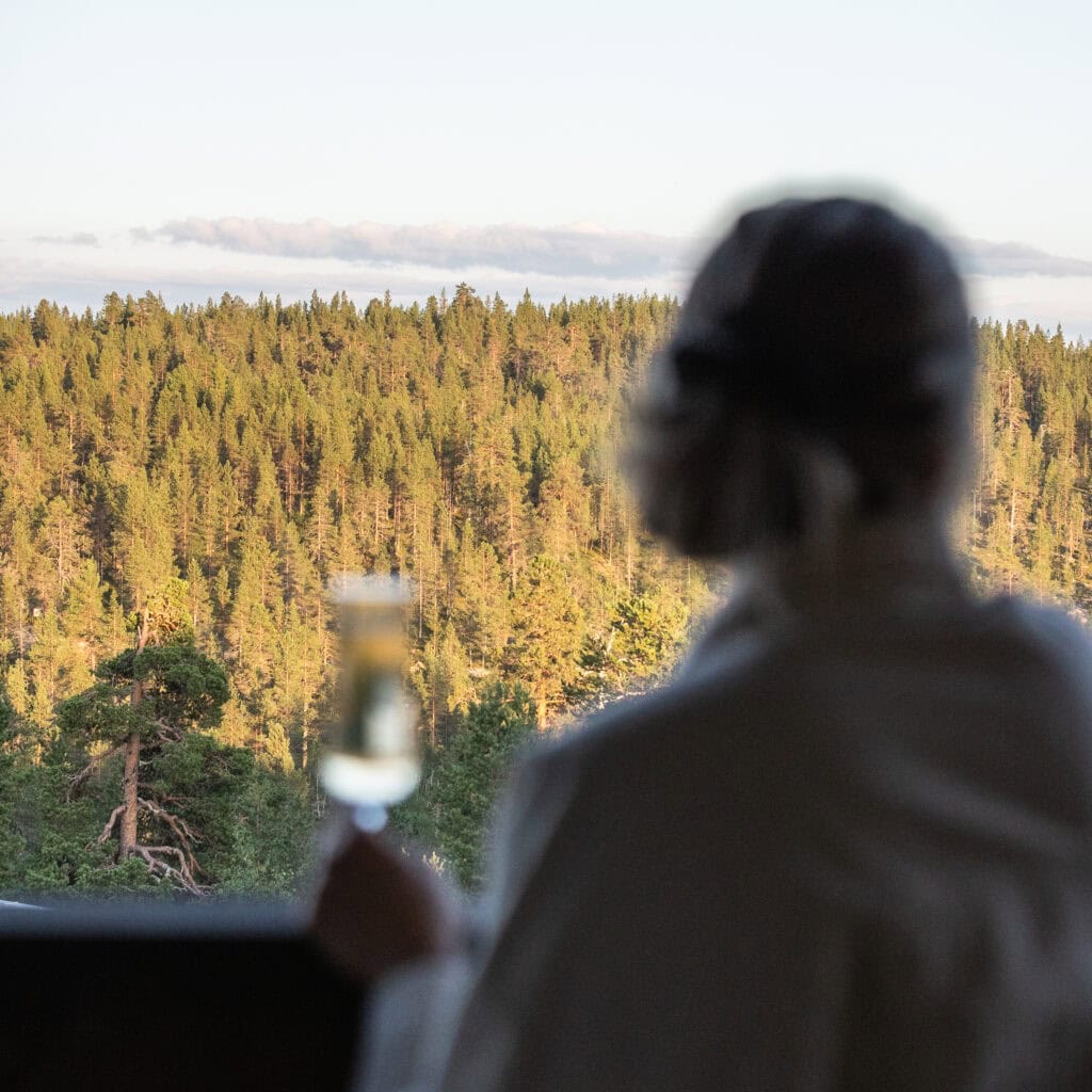 Nainen parvekkeella kädessään lasi kuohuvaa, taustalla metsämaisema