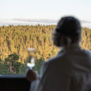 Nainen parvekkeella kädessään lasi kuohuvaa, taustalla metsämaisema